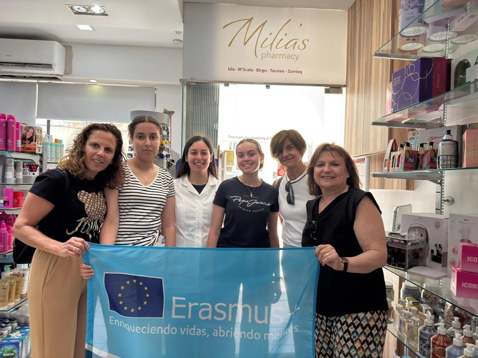 Students, teachers and a pharmacist holding an Erasmus+ flag inside a pharmacy